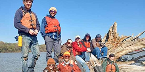 A group of students participating in a GIS field experience stand on, around and sit on a log.